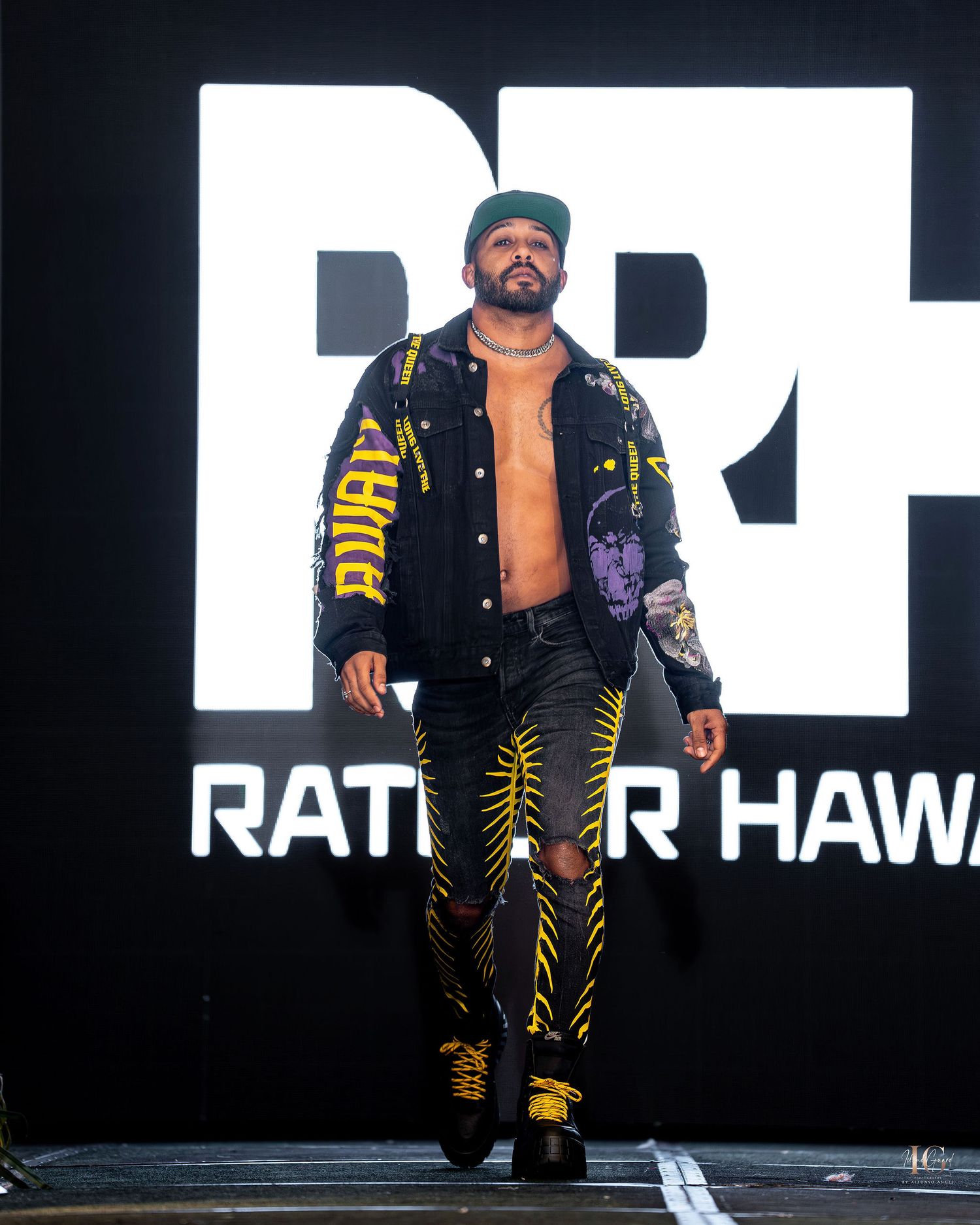 Professional wrestler in black and gold ring gear poses confidently against a dark background with text signage.