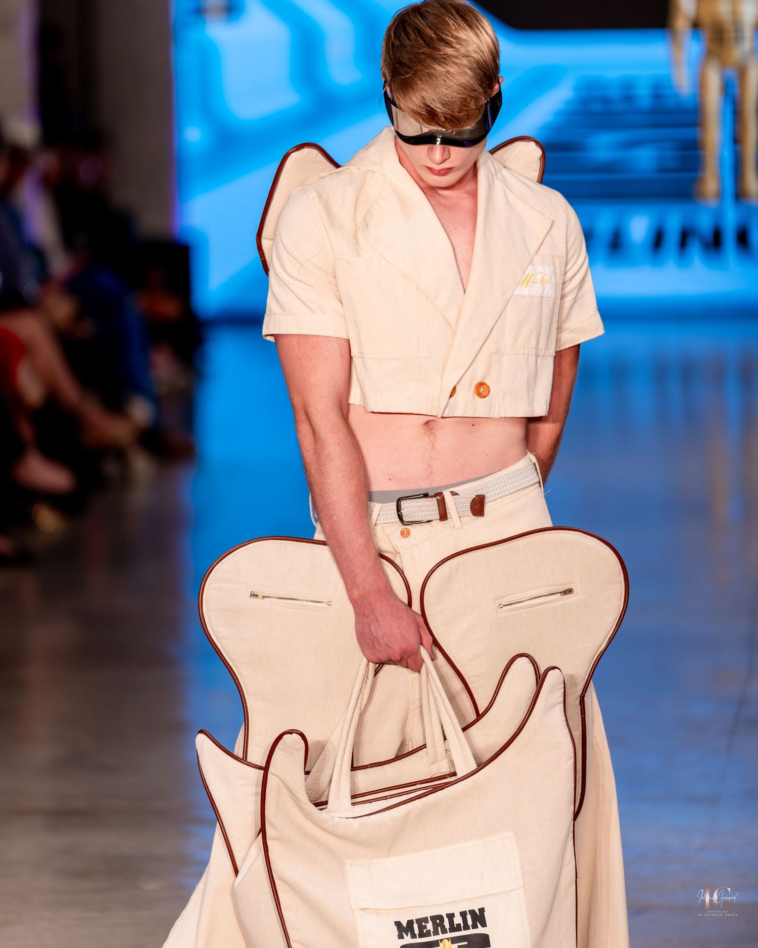 Fashion model walks runway in cream-colored utility dress with belt and pockets during fashion show presentation.