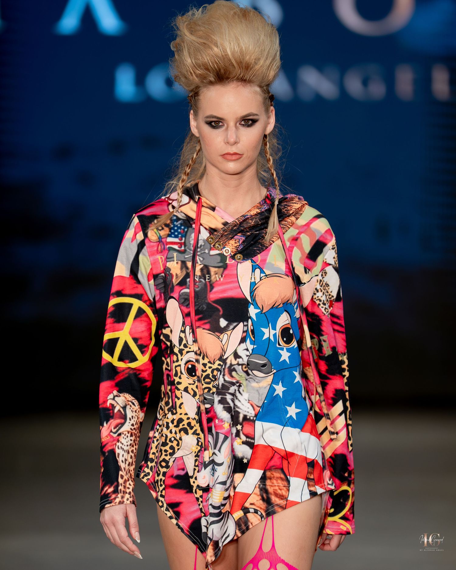 Model walks runway in colorful peace sign and American flag print jacket with dramatic upswept hairstyle at fashion show.