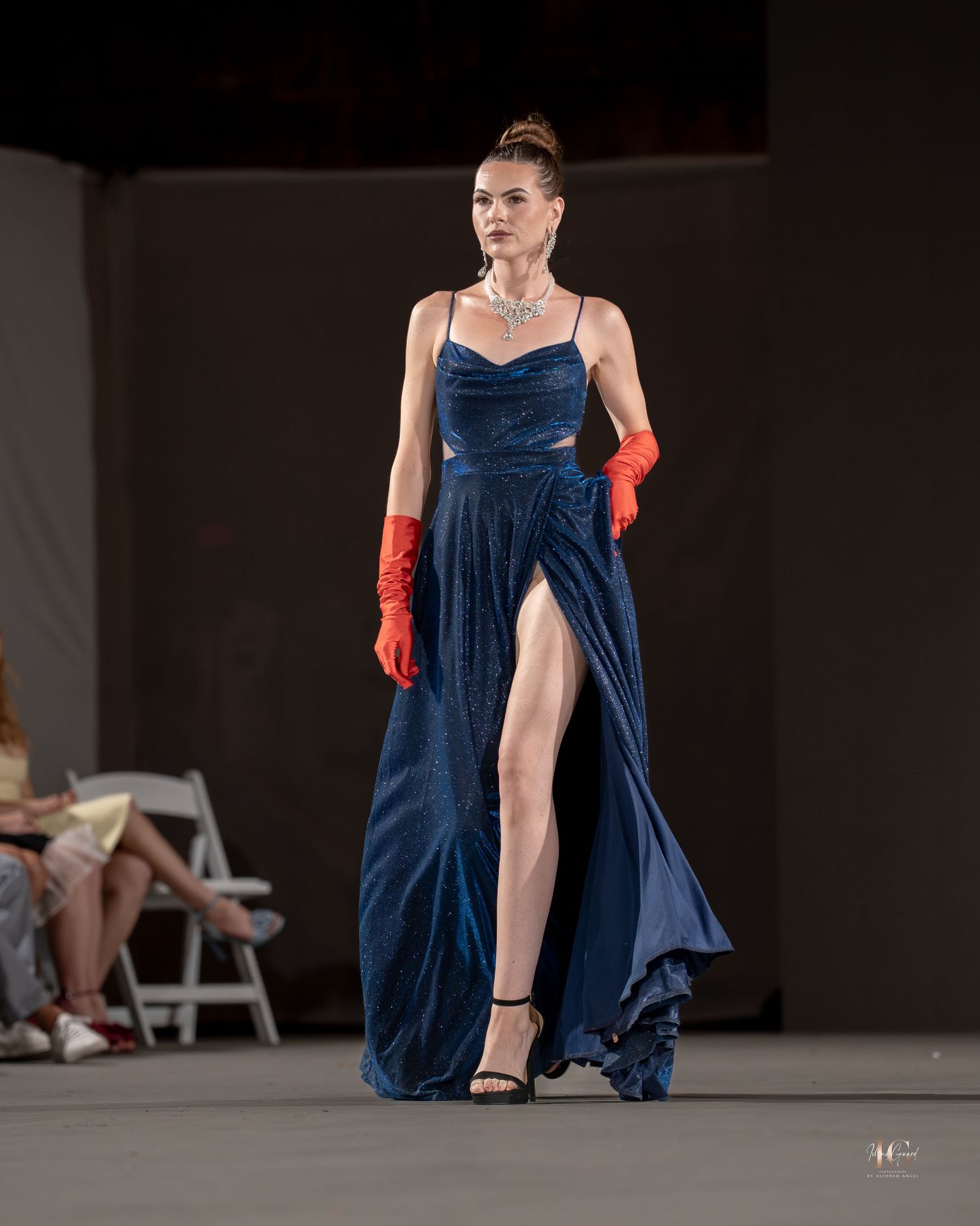 Model walks runway in flowing navy blue dress with red gloves during fashion show presentation.