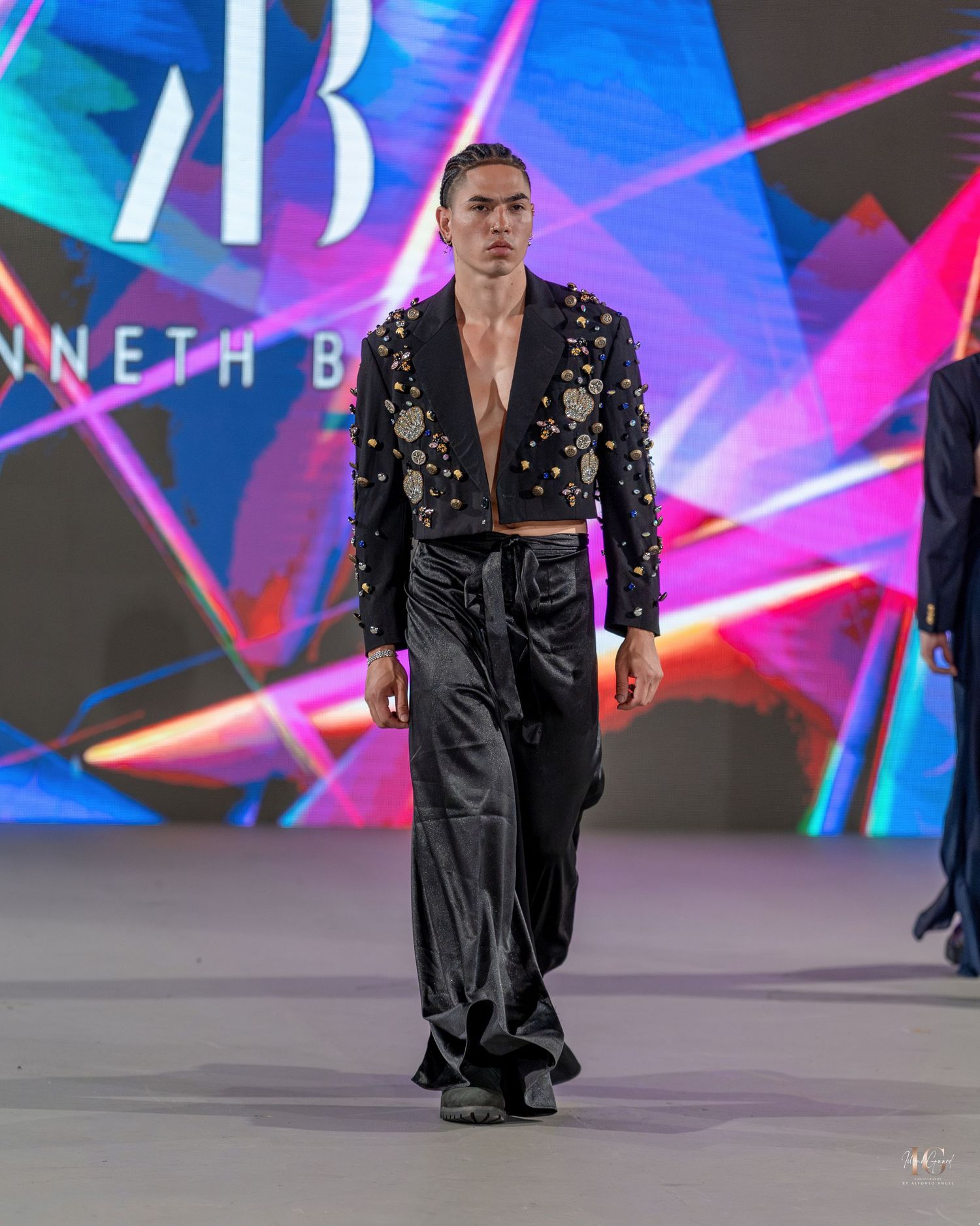 Fashion model walks runway in black embellished jacket and satin pants against vibrant neon backdrop at Kenneth B show.