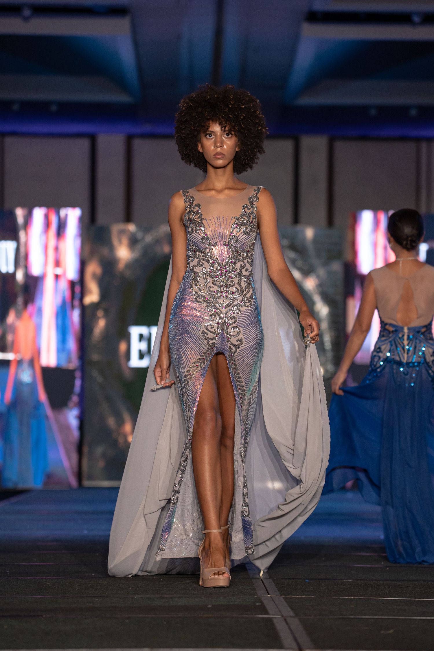 Model walks runway in sheer silver embellished gown with high slit and flowing cape detail at fashion show.