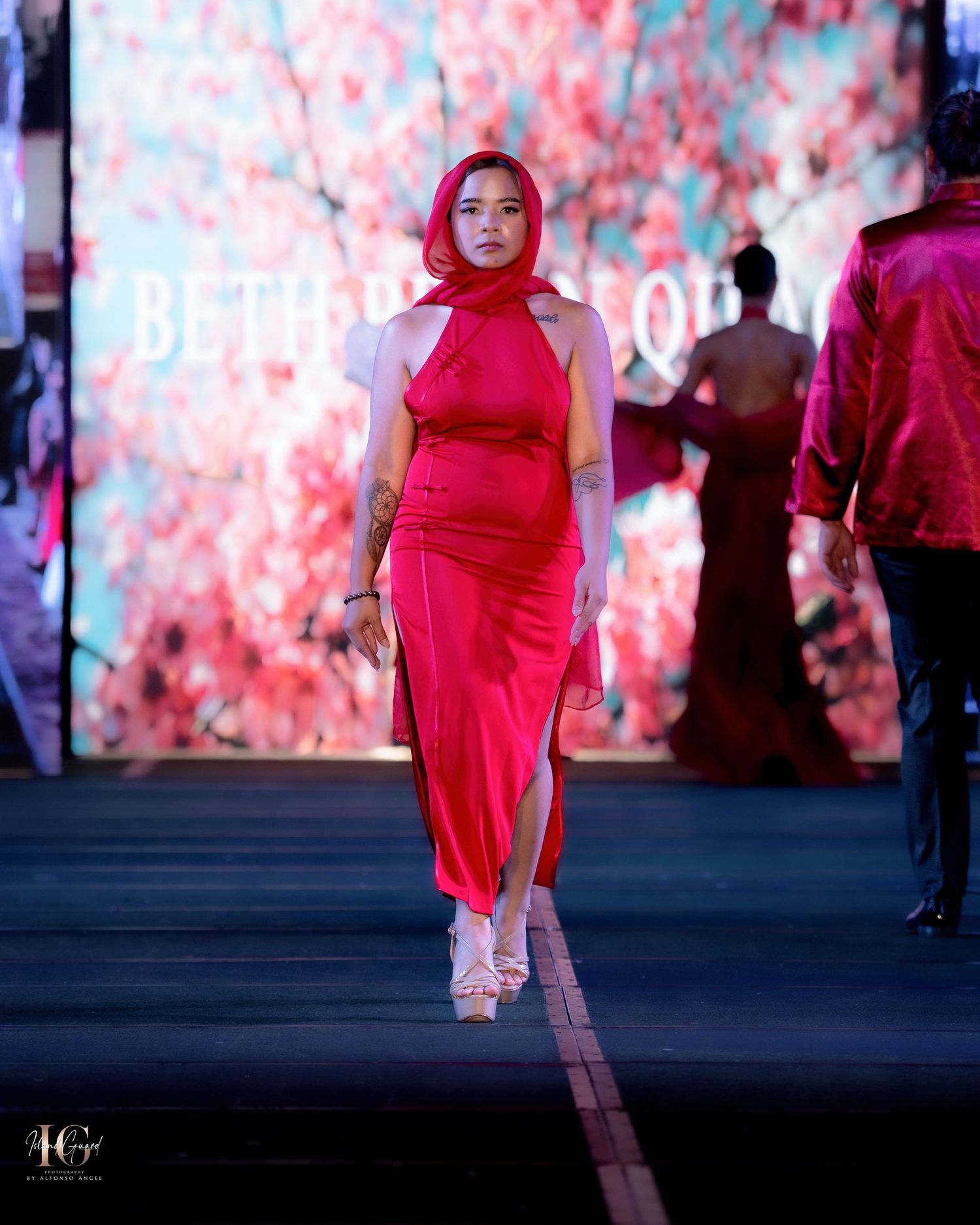 Model in sleek red dress walks runway against pink-lit backdrop.
