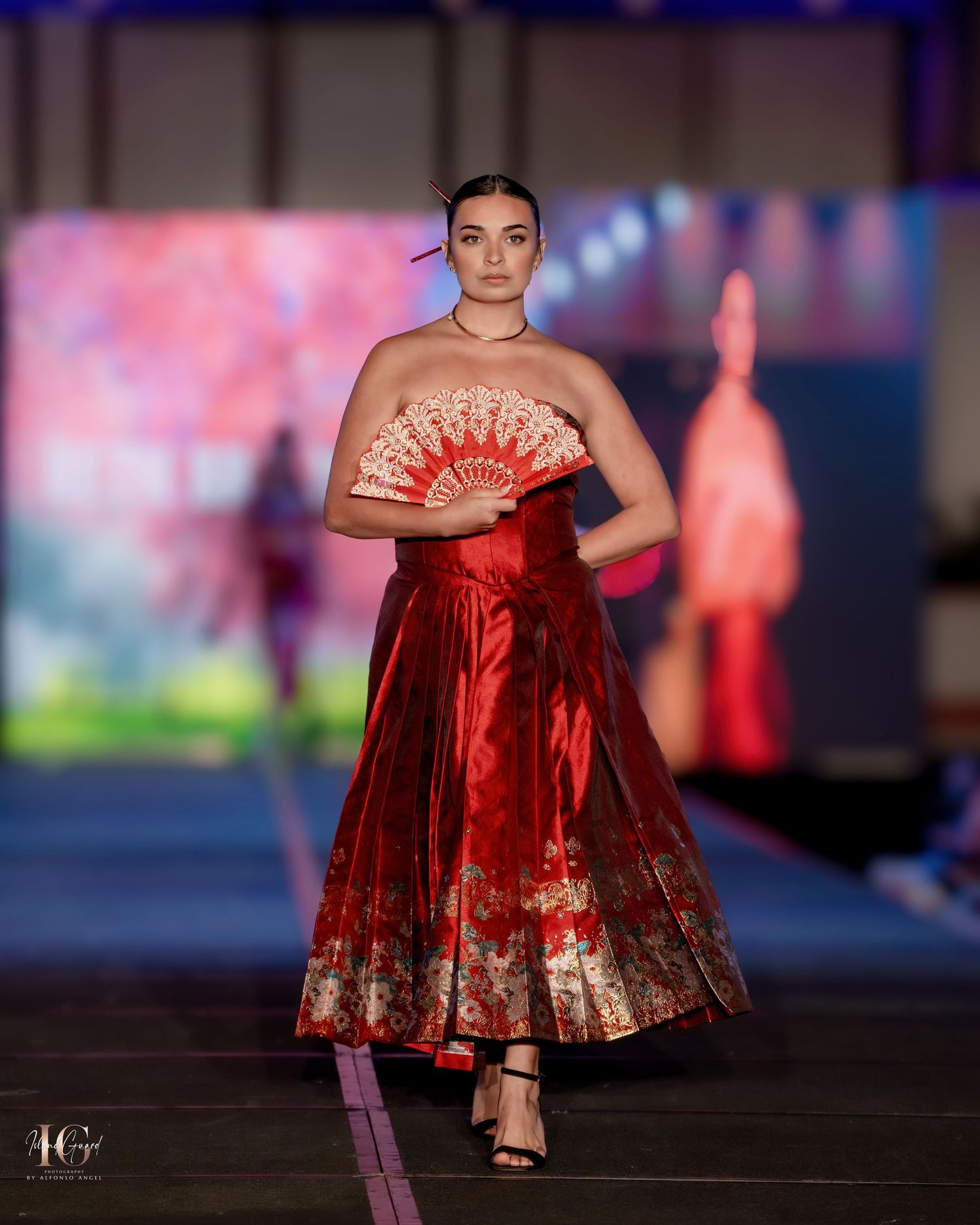 Model displays burgundy satin gown with decorative fan on illuminated catwalk.