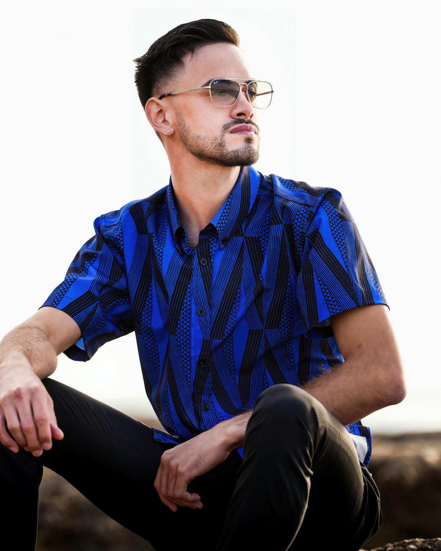 A man in glasses wearing a vibrant blue and black patterned polo shirt poses while kneeling outdoors.