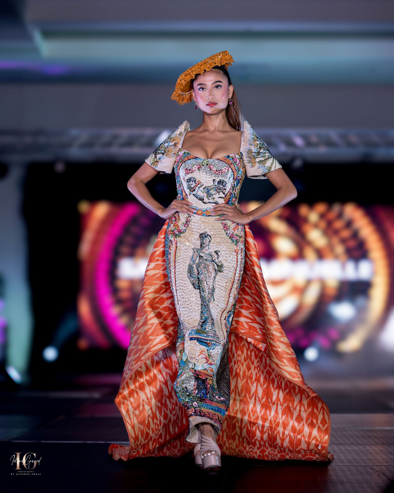 Fashion model walks runway in dramatic orange and silver dress with detailed embellishments at glamorous fashion show.