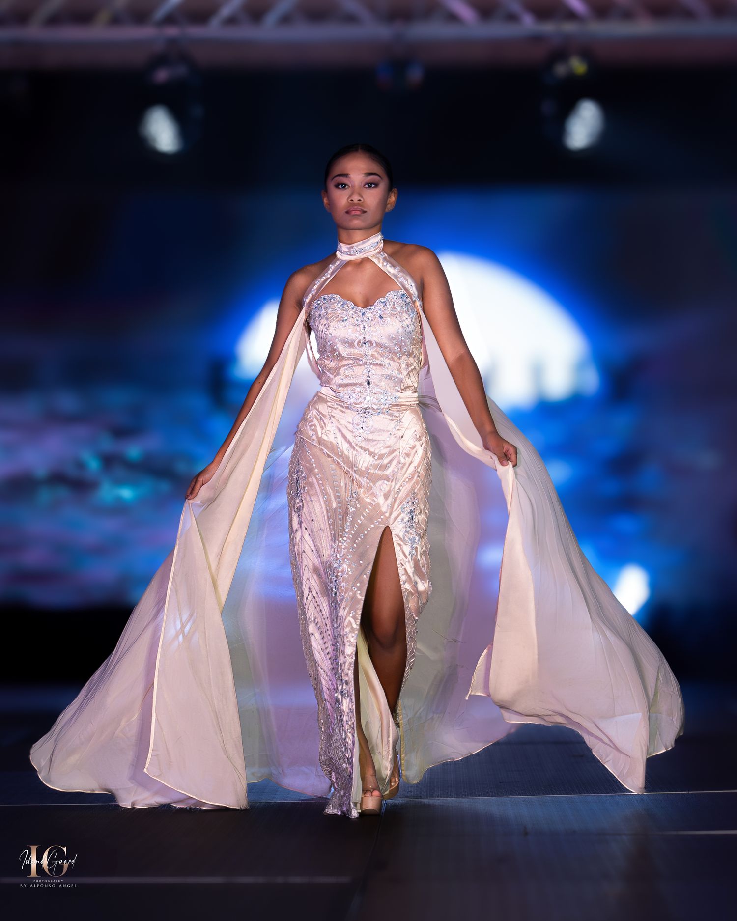 Elegant pink satin gown with flowing cape and halter neckline displayed on fashion runway.