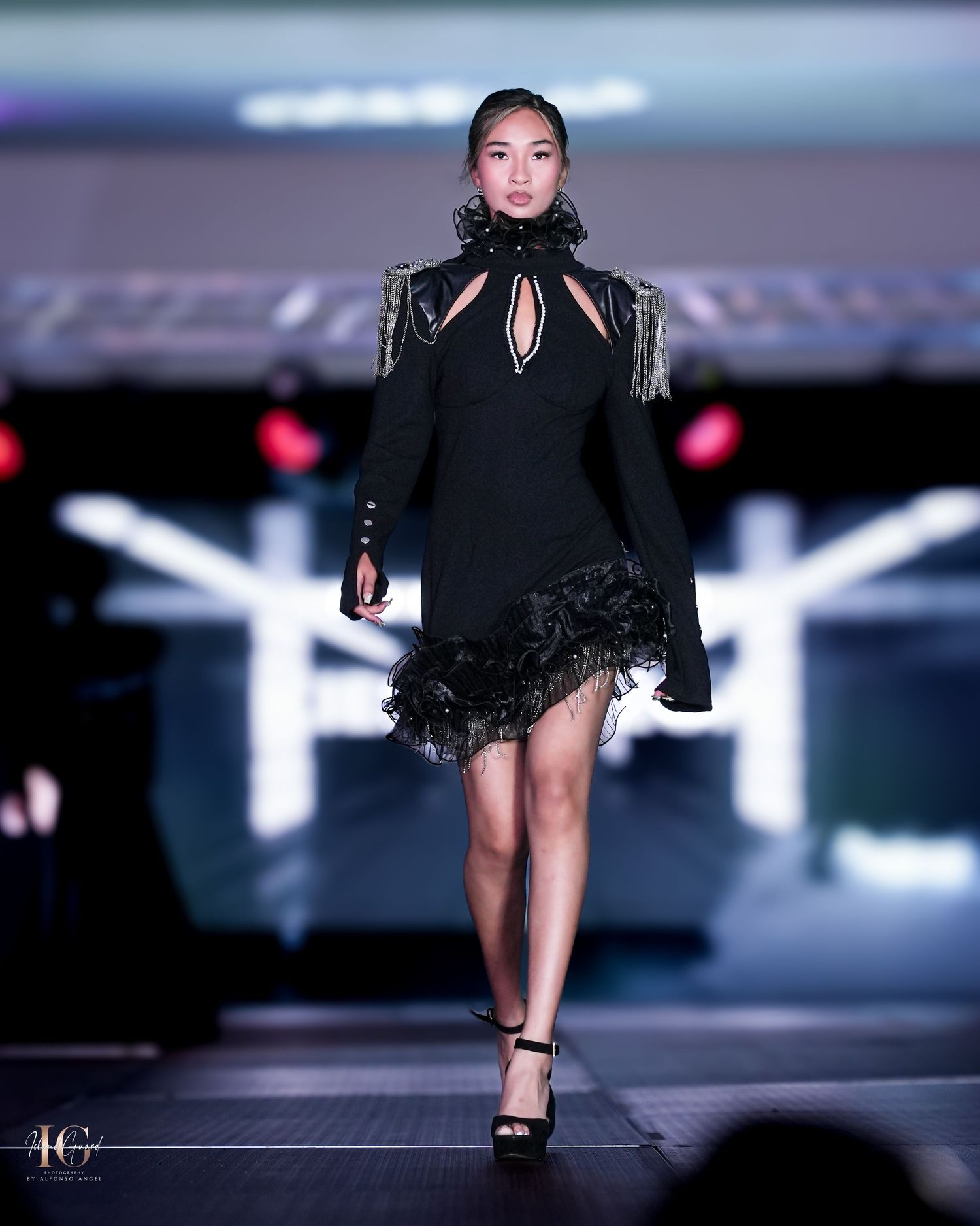 Model wearing elegant black mini dress with shoulder fringe and cutout details on fashion runway.