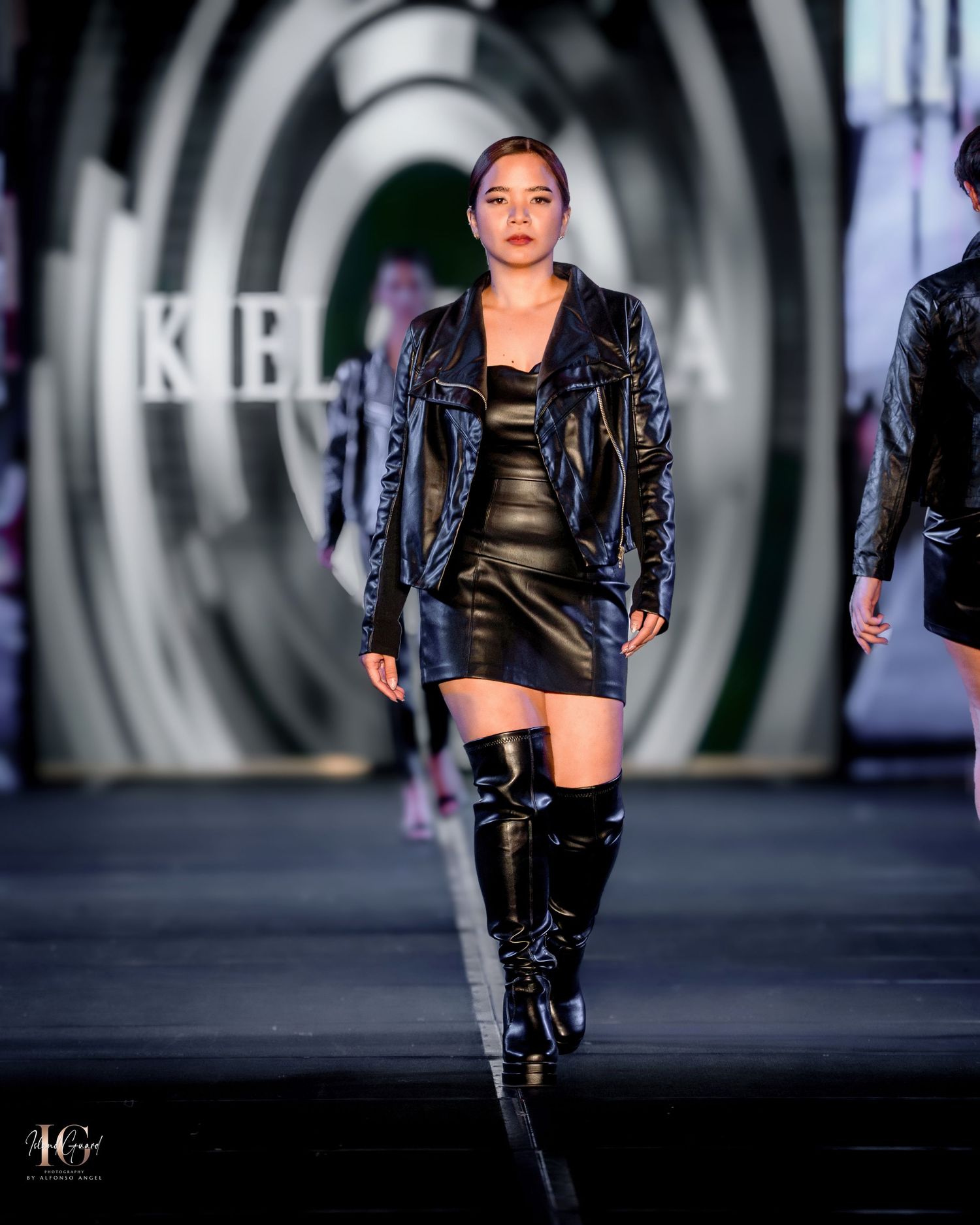 Model presents shiny leather mini dress with matching jacket and tall boots on catwalk.