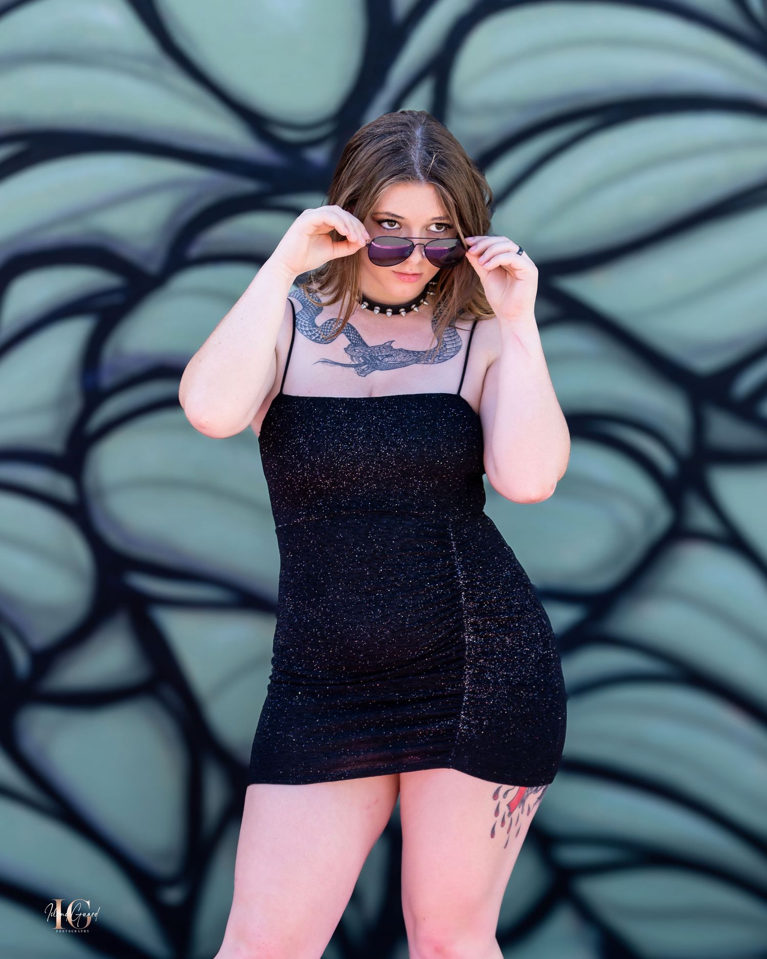 Fashion model in black sparkly dress playfully adjusts sunglasses against painted wall.