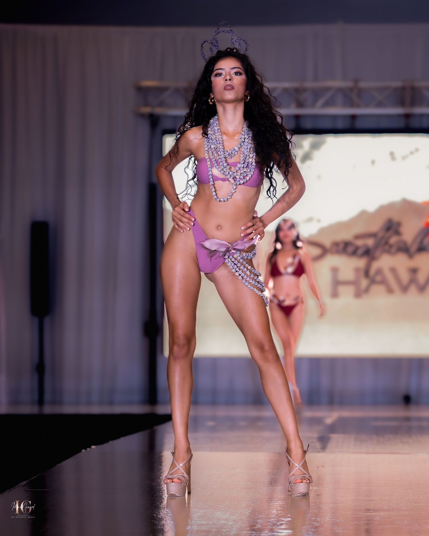 Model showcases embellished purple bikini on fashion runway with dramatic lighting.
