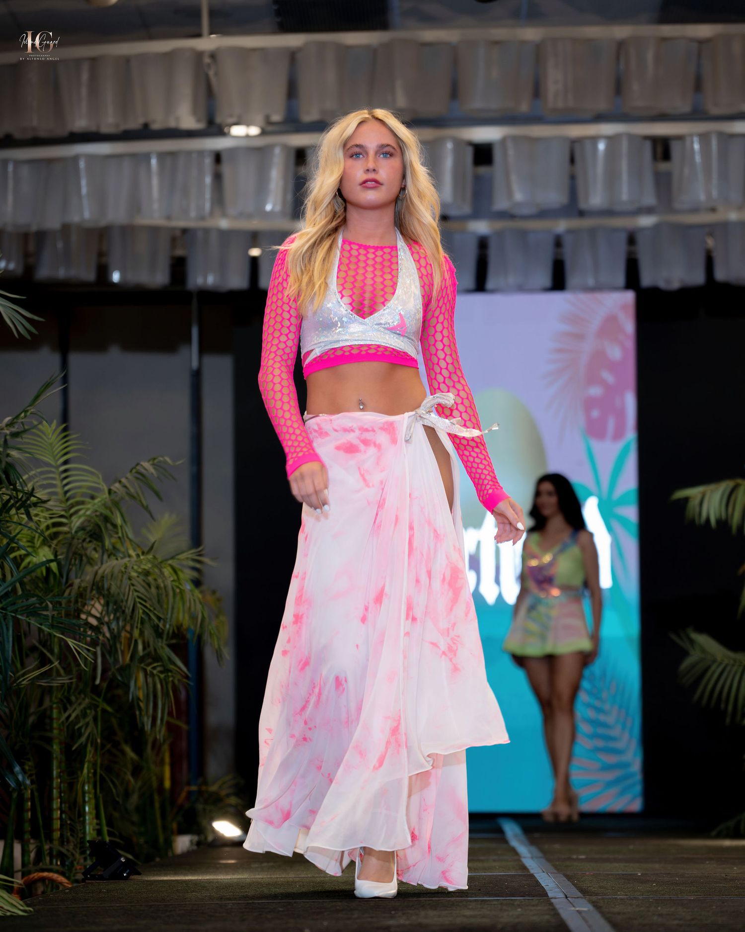 Model walks runway in pink crop top with matching sheer overlay and flowing white tie-dye maxi skirt during fashion show.