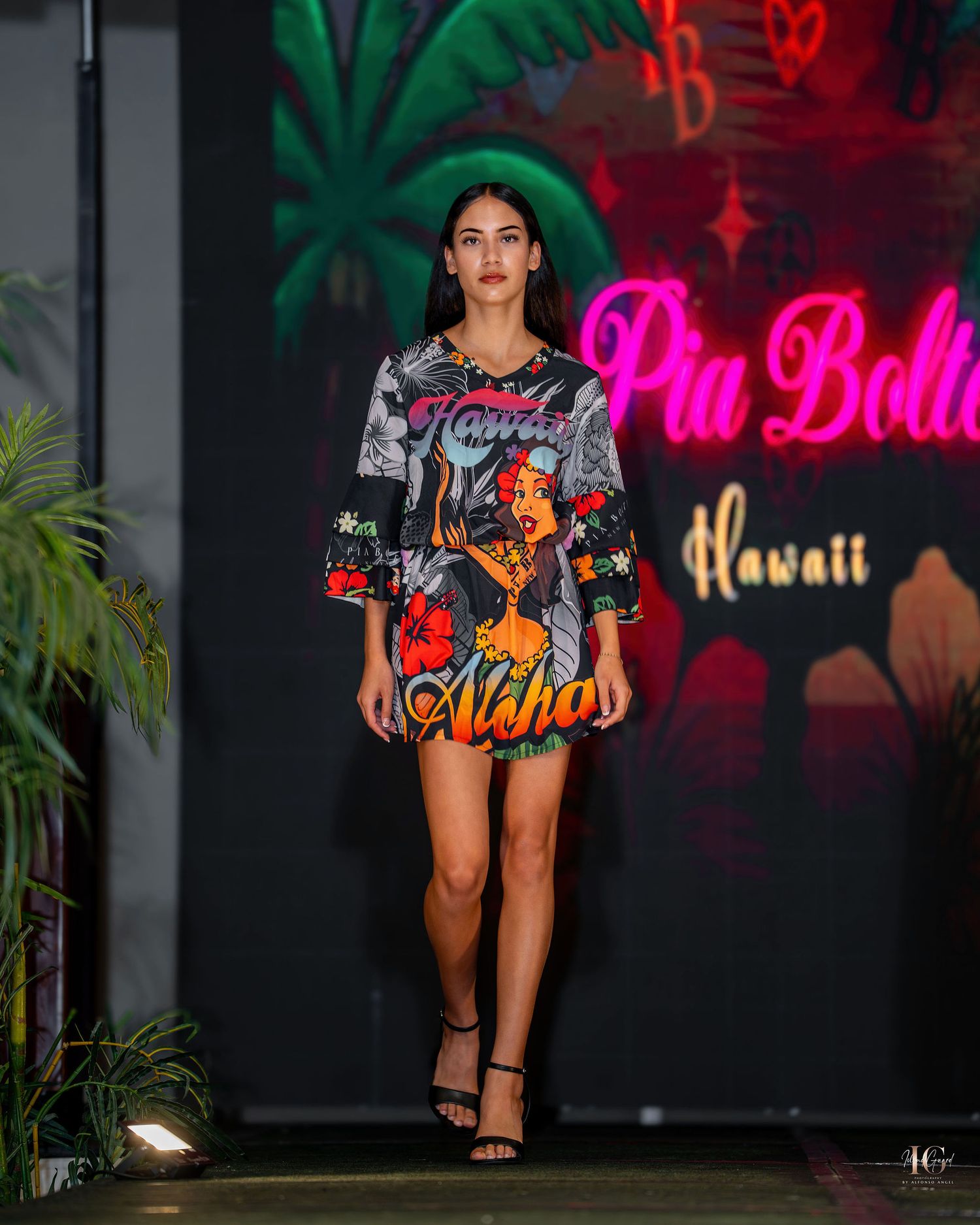 Model walks runway in colorful tropical print dress against neon-lit backdrop at fashion show.