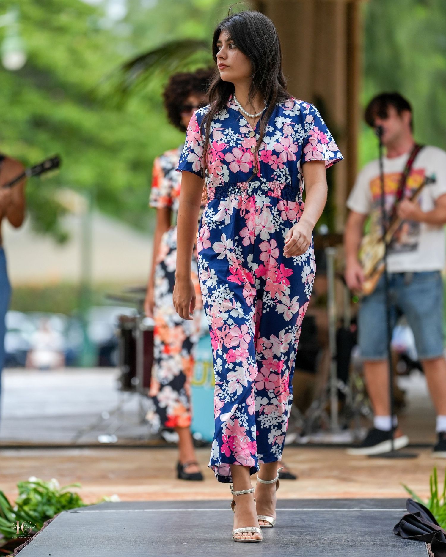 Person wearing a colorful blue and pink floral dress walks on a sidewalk near trees and parked cars.