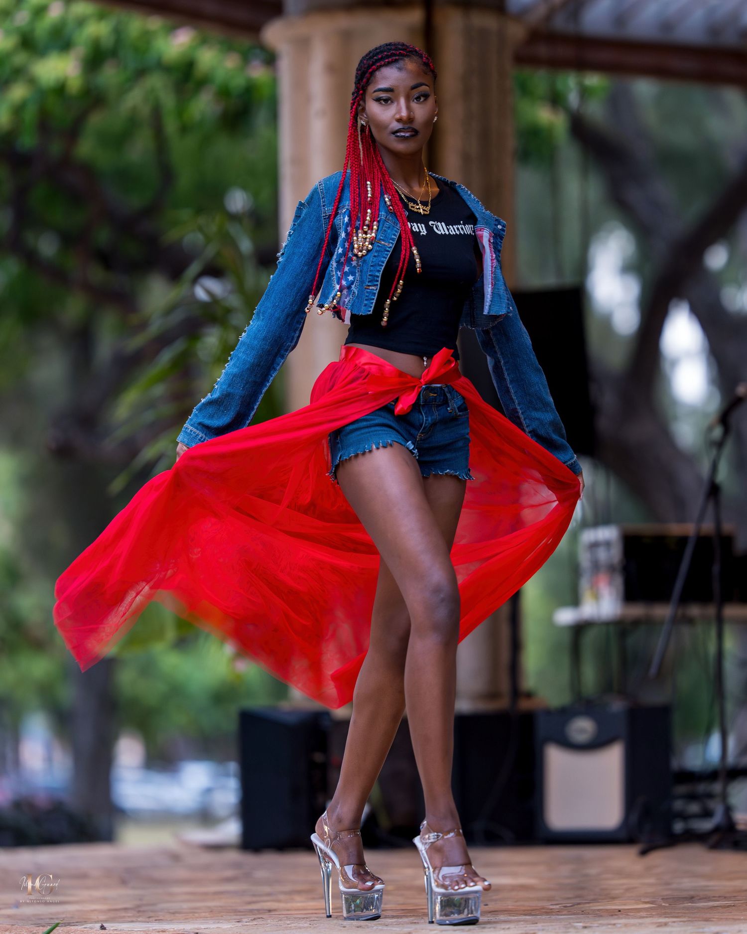 Model poses in denim jacket and shorts with flowing red skirt overlay and platform heels during outdoor photoshoot.