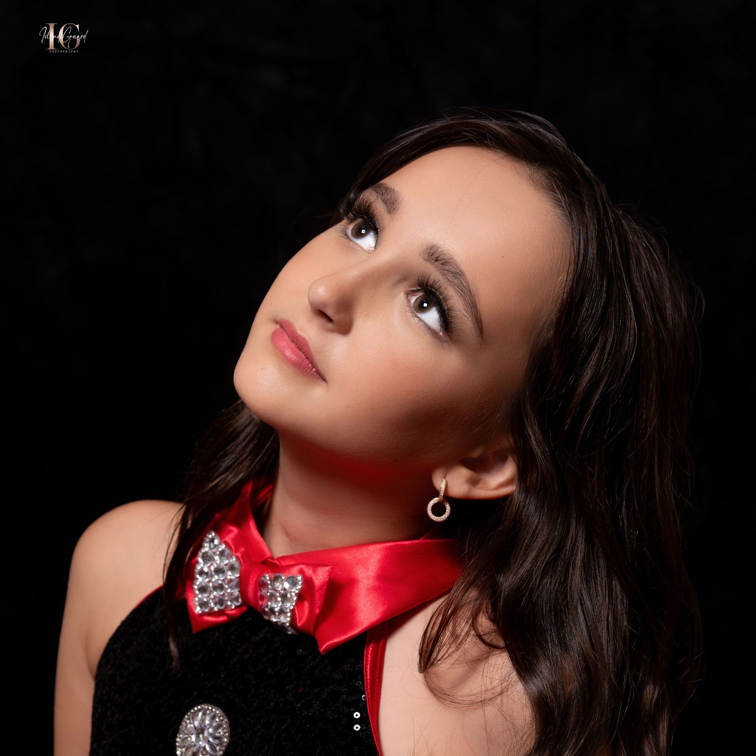 Portrait of someone with dark hair wearing black attire and a red satin collar with crystal accents looking upward.