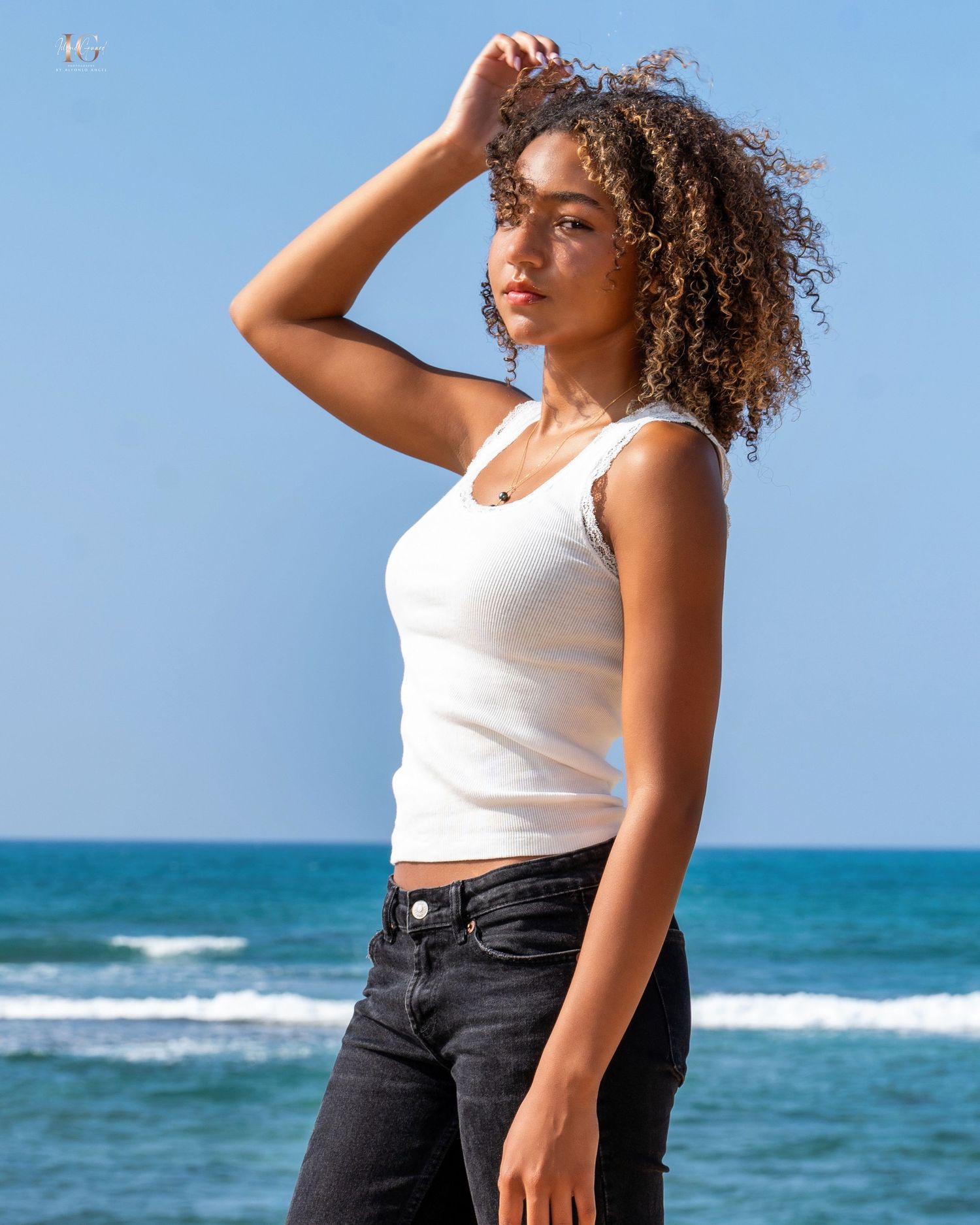 Person in white tank top and dark jeans posing against blue ocean backdrop on sunny day.