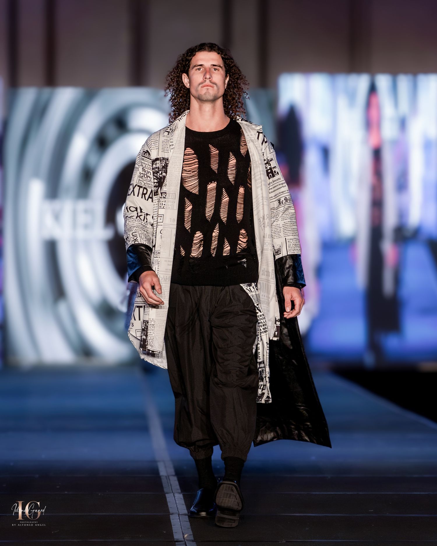 Model presents newspaper-print jacket with black ensemble on modern fashion runway.