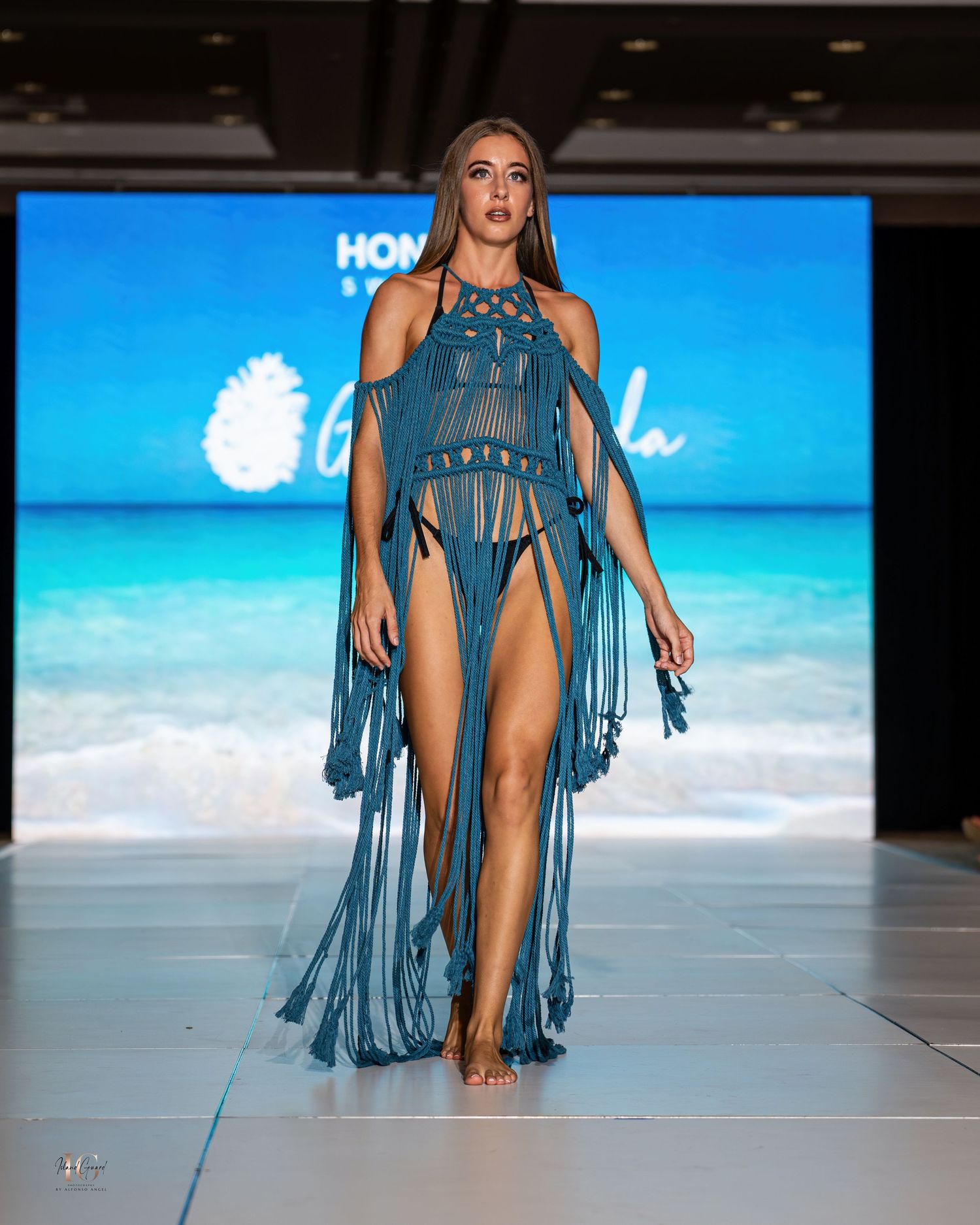 Blue fringe swimwear design showcased on runway with tropical beach background.