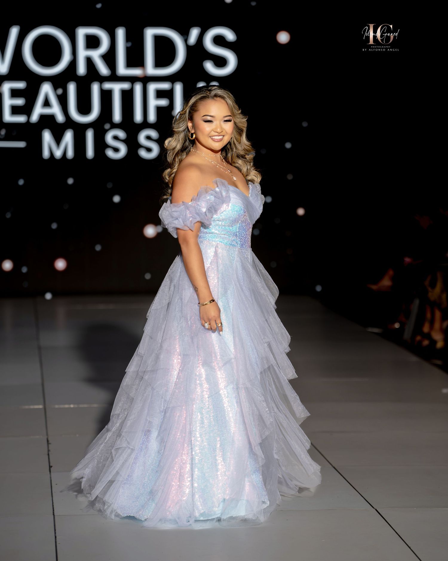 Model walks runway in iridescent light blue tulle gown with off-shoulder design during fashion show.
