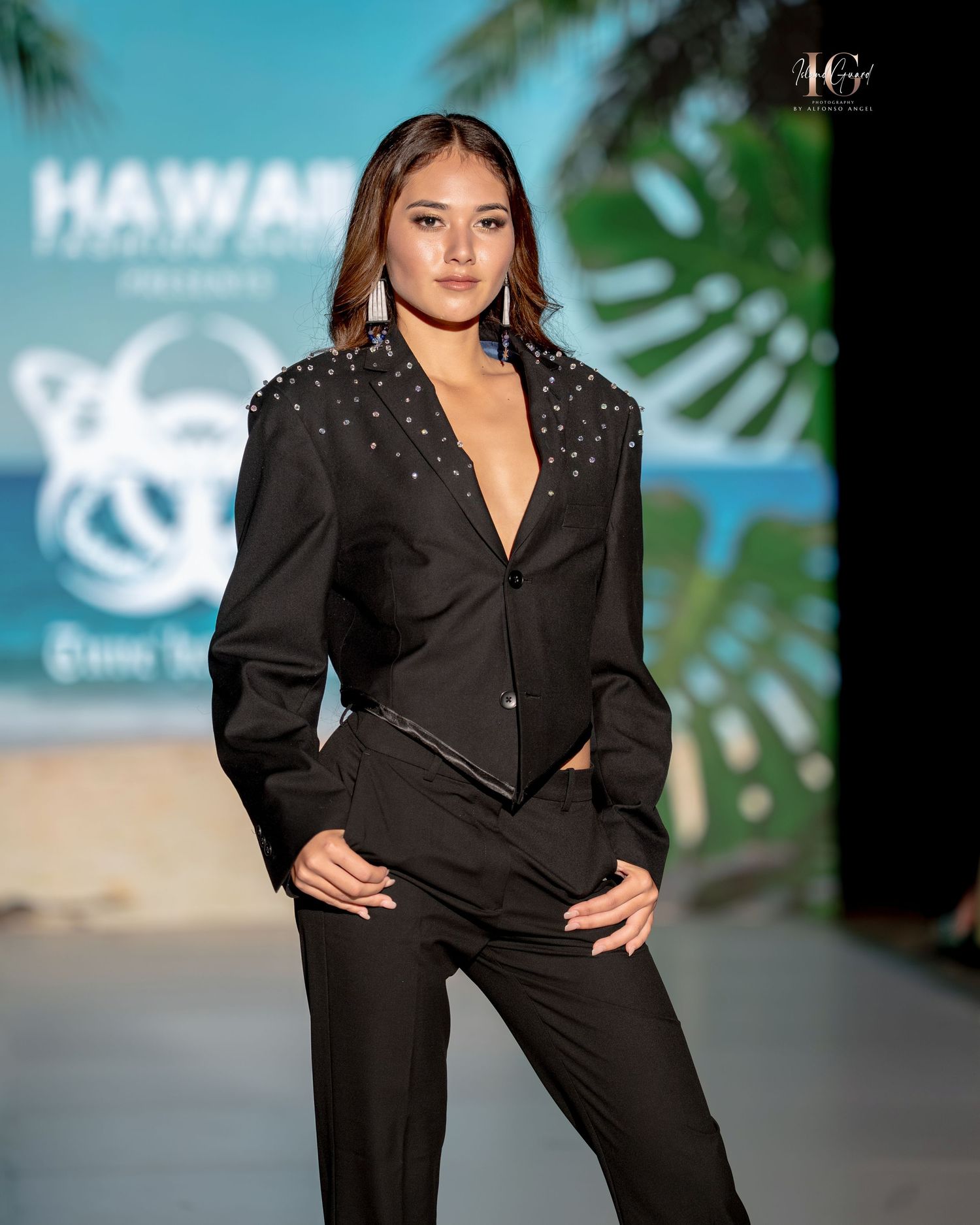Model walks runway in black structured blazer and pants during fashion show with tropical backdrop.