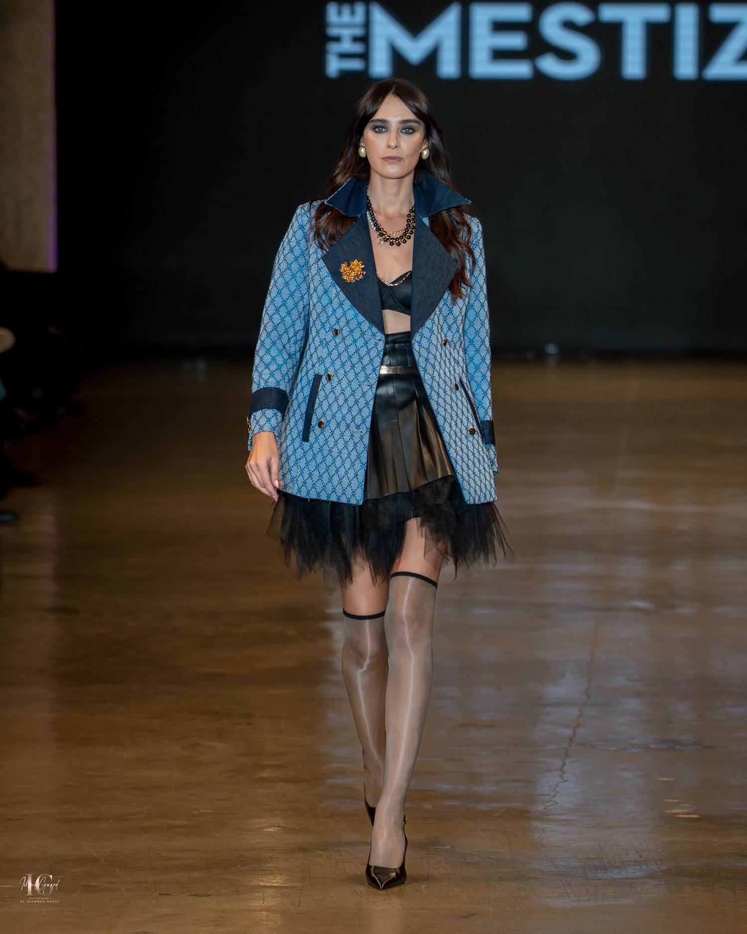 Model walks runway in light blue patterned blazer and black dress during fashion show.