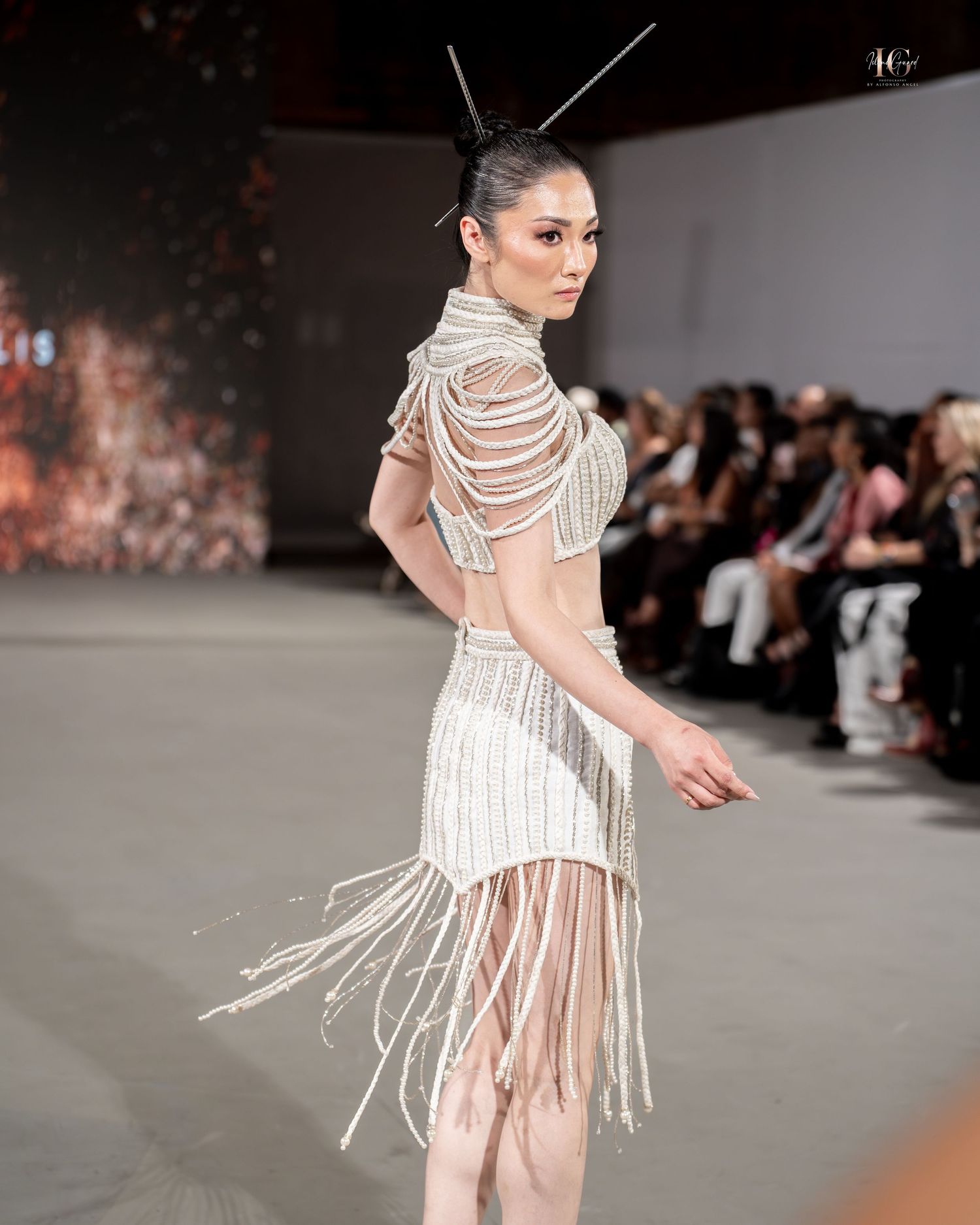 Model walks runway in sheer white dress with dramatic pearl fringe detailing at fashion show.