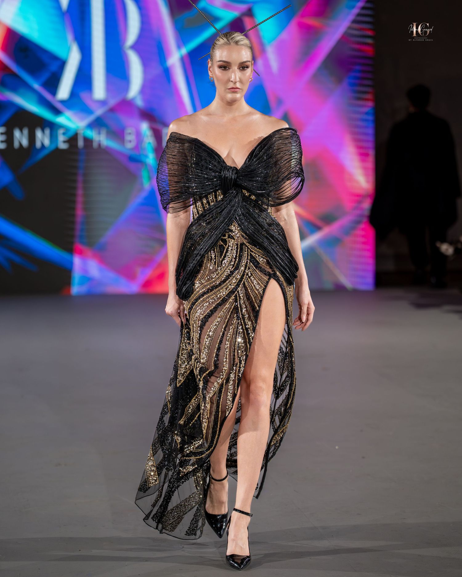 A model walks the runway in a black sequined evening gown with high slit and off-shoulder design at a fashion show.