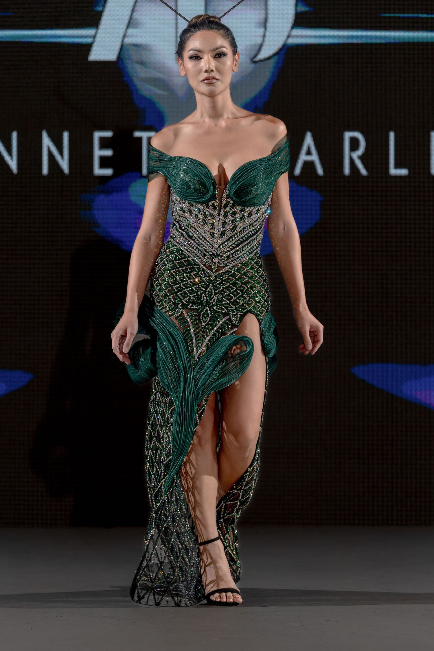 A model walks the runway in an emerald green beaded evening gown with a high slit and intricate embellishments.