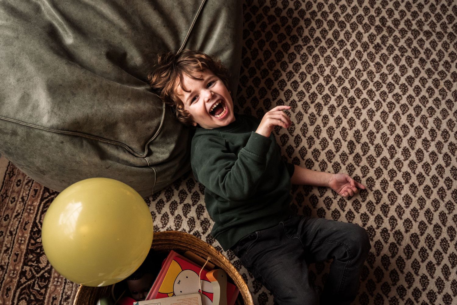 Child lying on patterned carpet with a yellow balloon and toys.