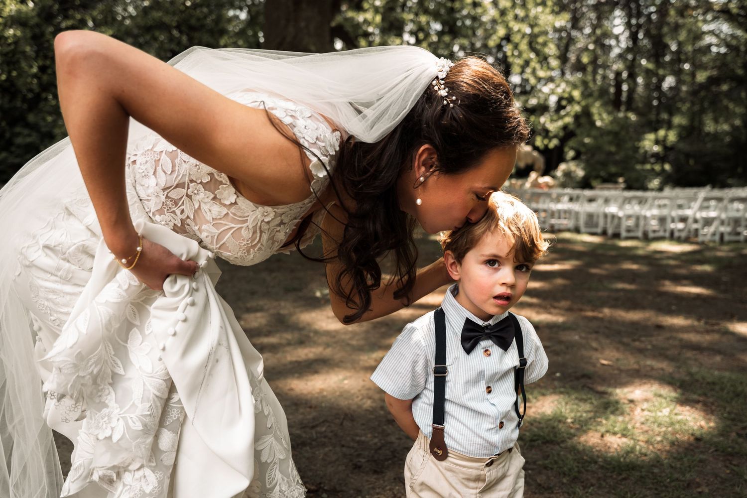A person in a white lace wedding dress bends down to help a small child wearing a white outfit in an outdoor setting.