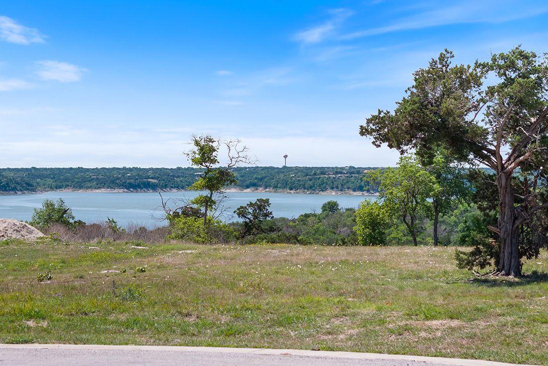 Scenic lakeside view with lush green trees, open grassland, and calm blue water under a partly cloudy sky.