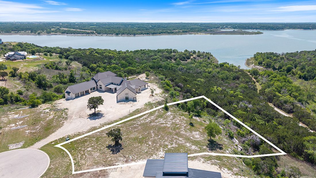 Aerial view of a lakeside property with a modern home, circular driveway, and outlined lot surrounded by lush trees.