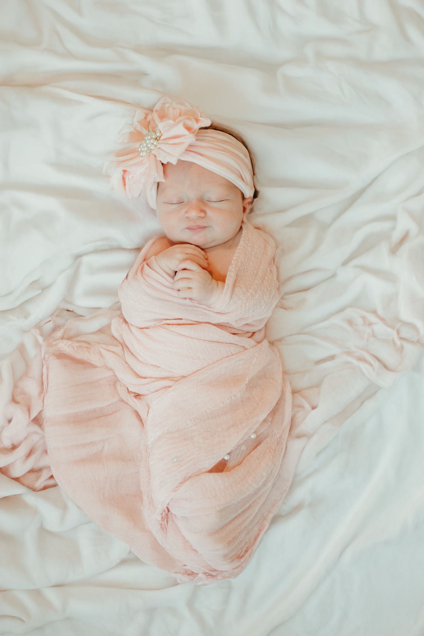 Peaceful newborn baby in pink swaddle and floral headband sleeping on white sheets.
