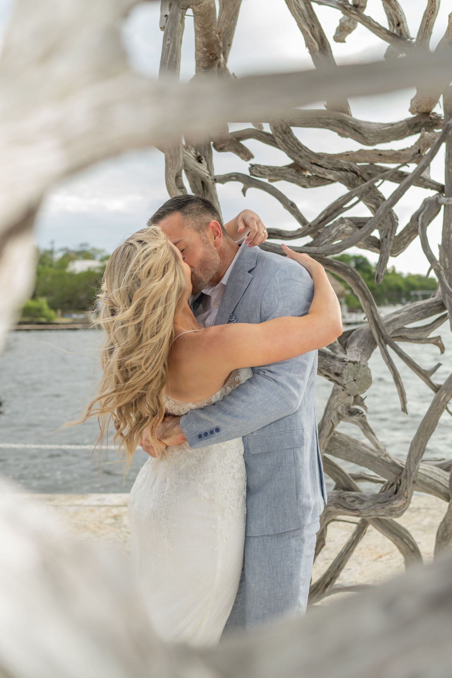 Romantic moment captured through driftwood branches at a waterfront wedding venue.