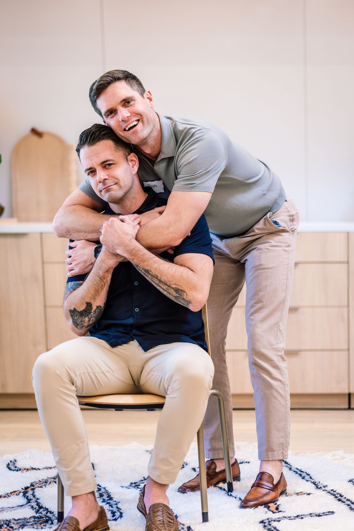 Two men share a warm embrace, one seated on a chair while the other stands behind him hugging him in a bright modern home.