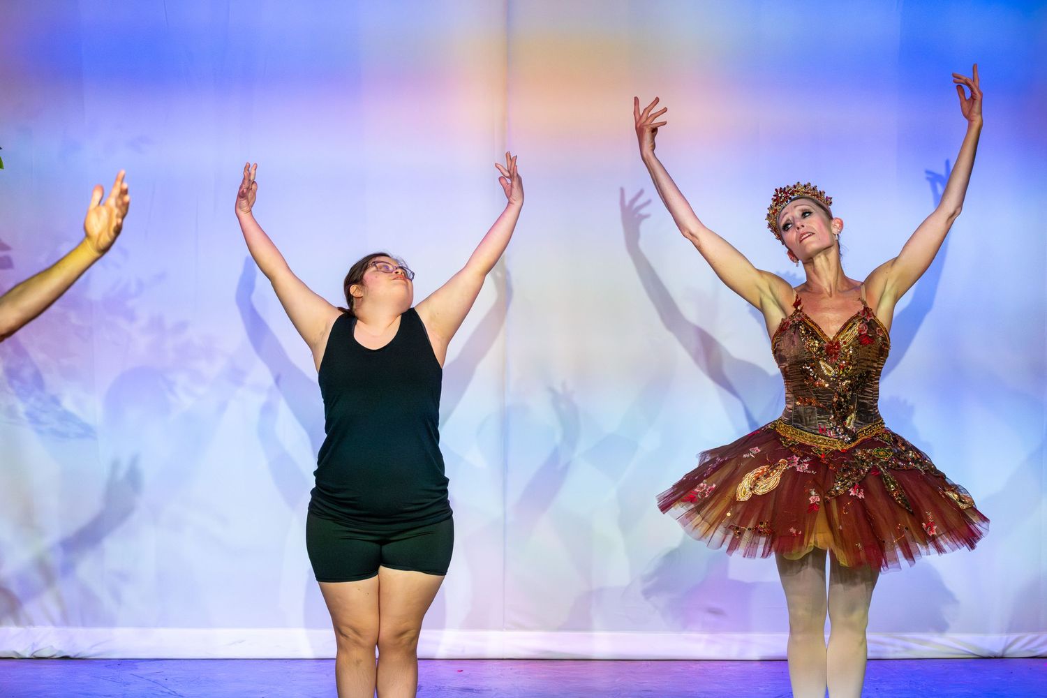 Two women perform on stage, one in a black outfit and one in a ornate ballet tutu, both with arms raised gracefully.