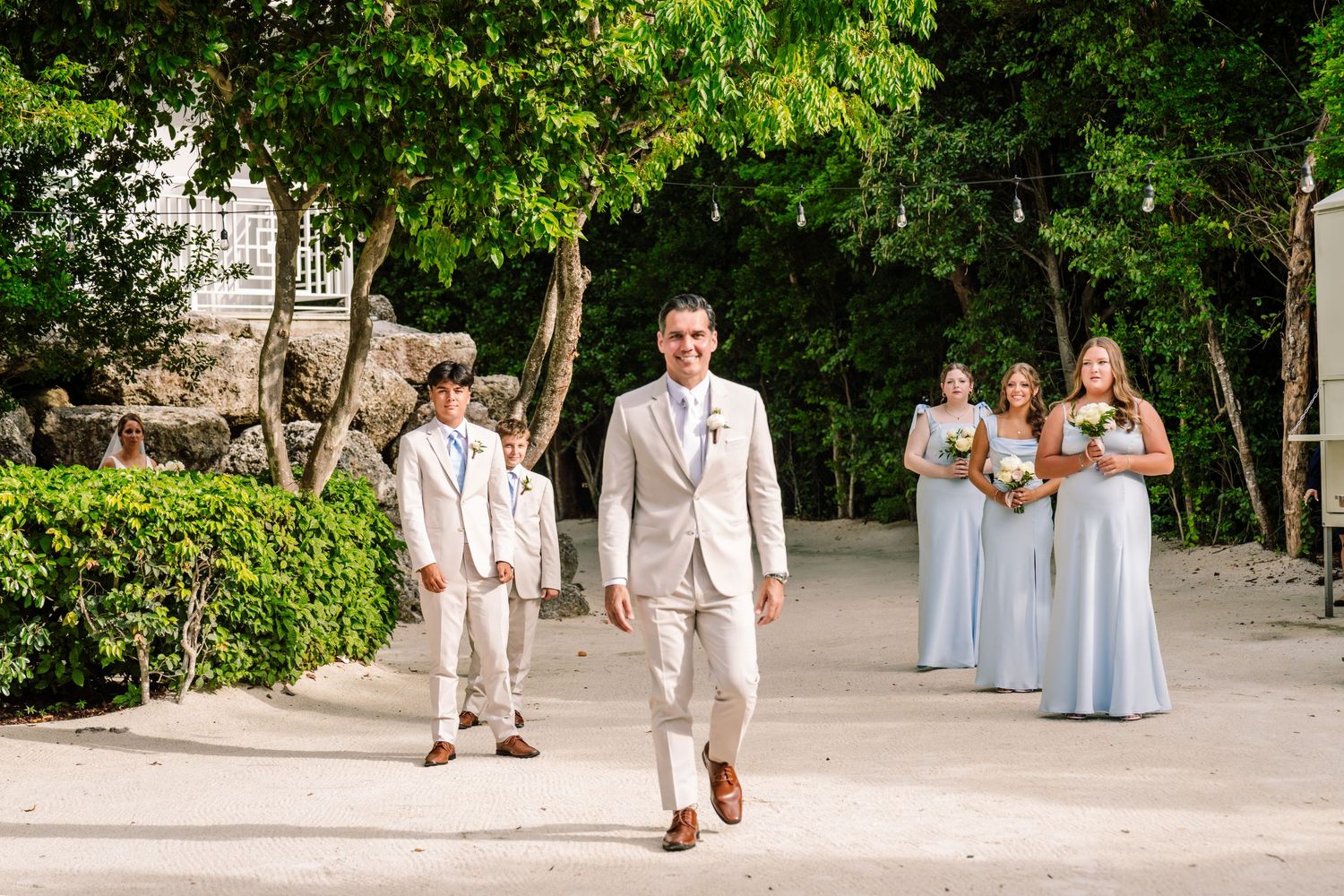 Wedding party in light colored attire walks along a garden path during an outdoor celebration.