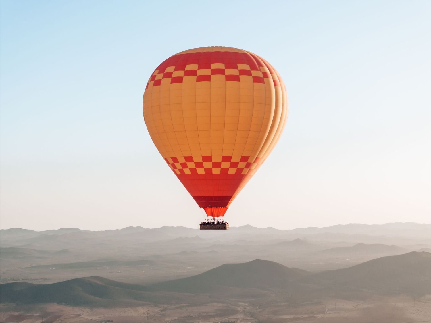 An orange and red checkered hot air balloon floats above misty mountain peaks at sunrise.