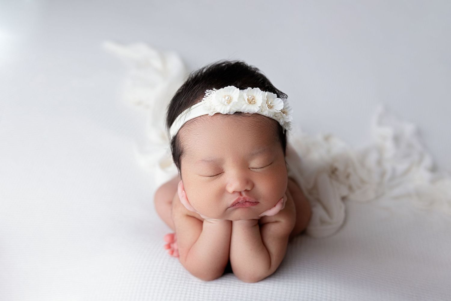 Sleeping newborn baby wearing white lace headband poses with hands under chin on fluffy white backdrop.