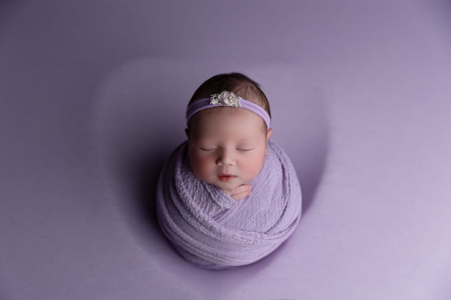 Sleeping newborn wrapped in soft purple swaddle on matching purple backdrop with delicate headband accessory.