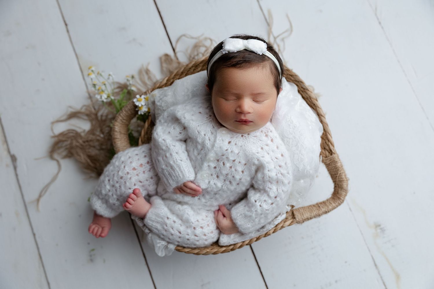 Sleeping newborn wearing white knit outfit nestled in rustic basket with floral decorations on white wooden floor.
