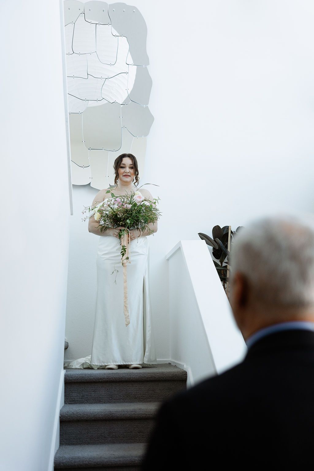 A bride in a white dress holds a floral bouquet while standing on a platform during an indoor ceremony.