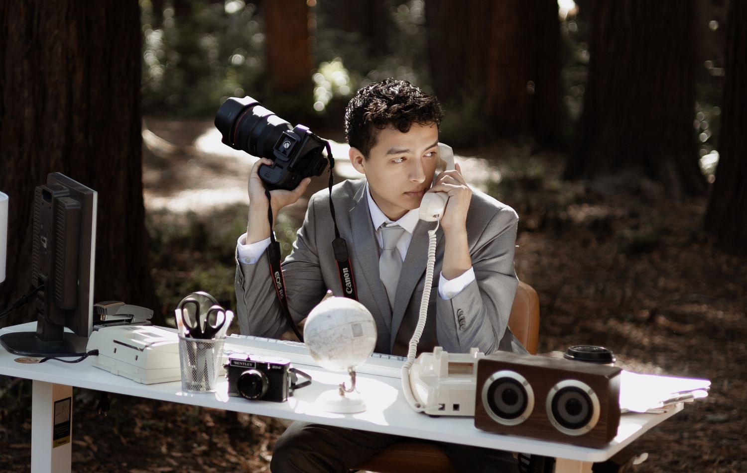 A photographer in a gray suit works at an outdoor desk setup with cameras and equipment in a forest setting.