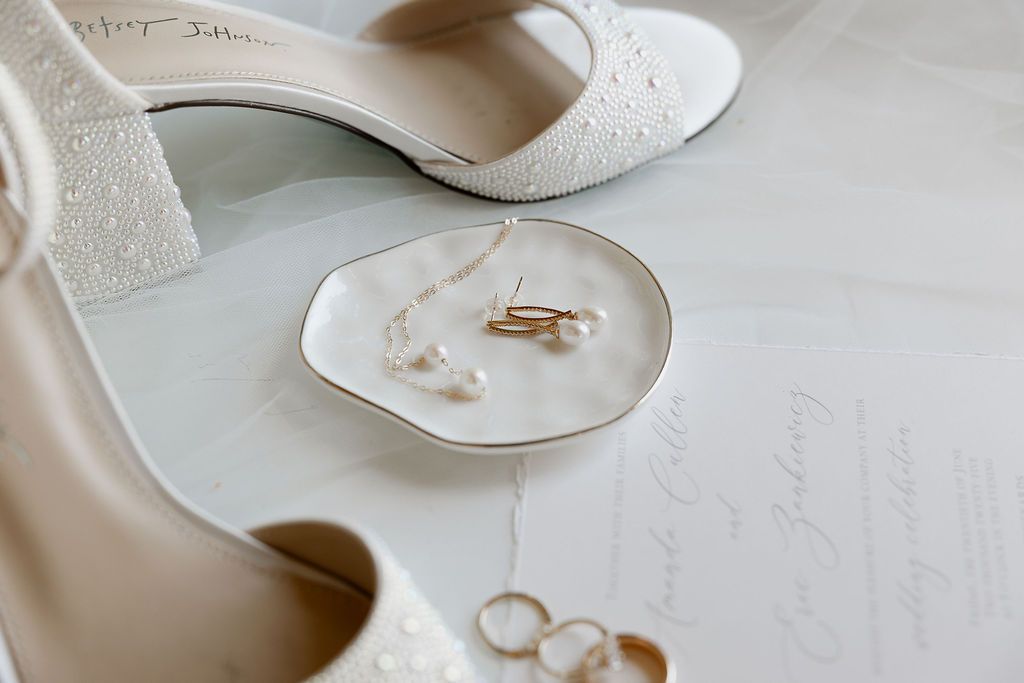 Elegant white bridal pumps and wedding rings arranged on neutral background for wedding detail photography.