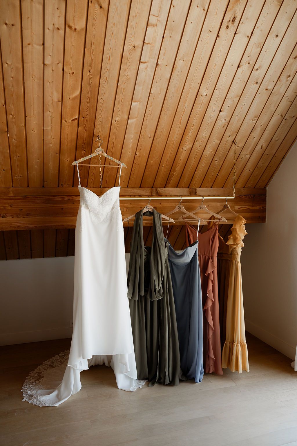 Wedding dresses and bridesmaid gowns hanging from a wooden beam in rustic attic space.