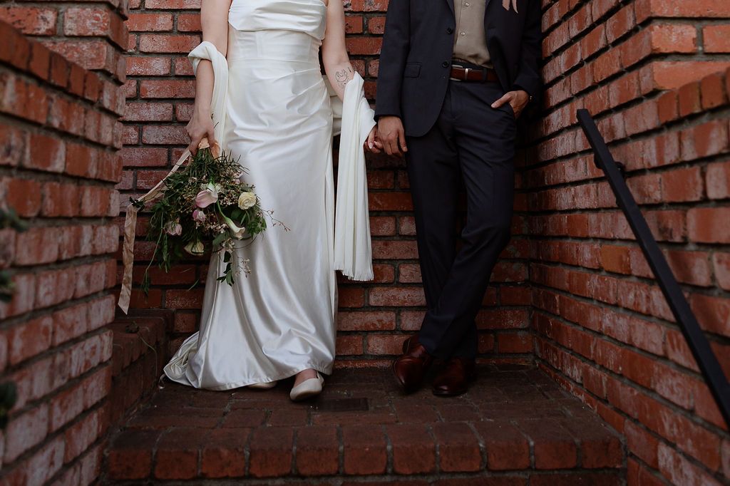 A couple in wedding attire poses against a rustic brick wall background creating romantic wedding portraits.