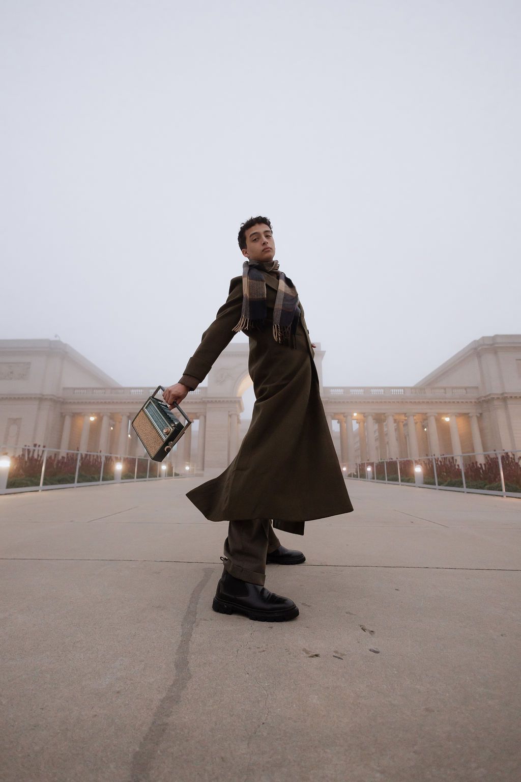 A person in a long brown coat strikes a dramatic pose outside a classical building in foggy weather, holding a bag.