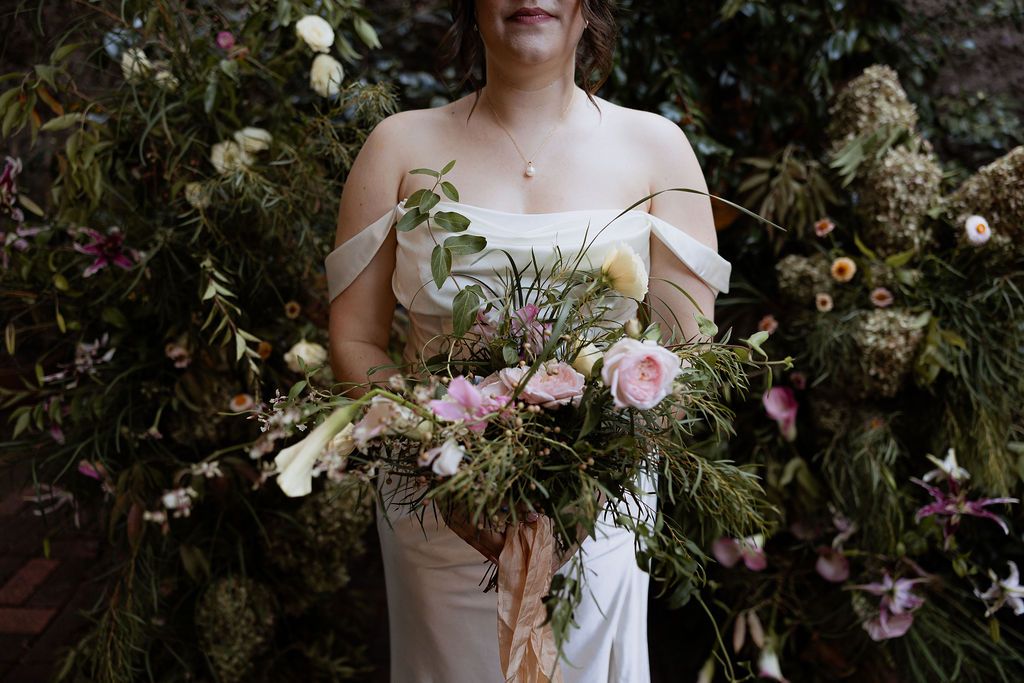 A romantic garden wedding bouquet with soft white and pink flowers sitting against a white dress in an outdoor setting.