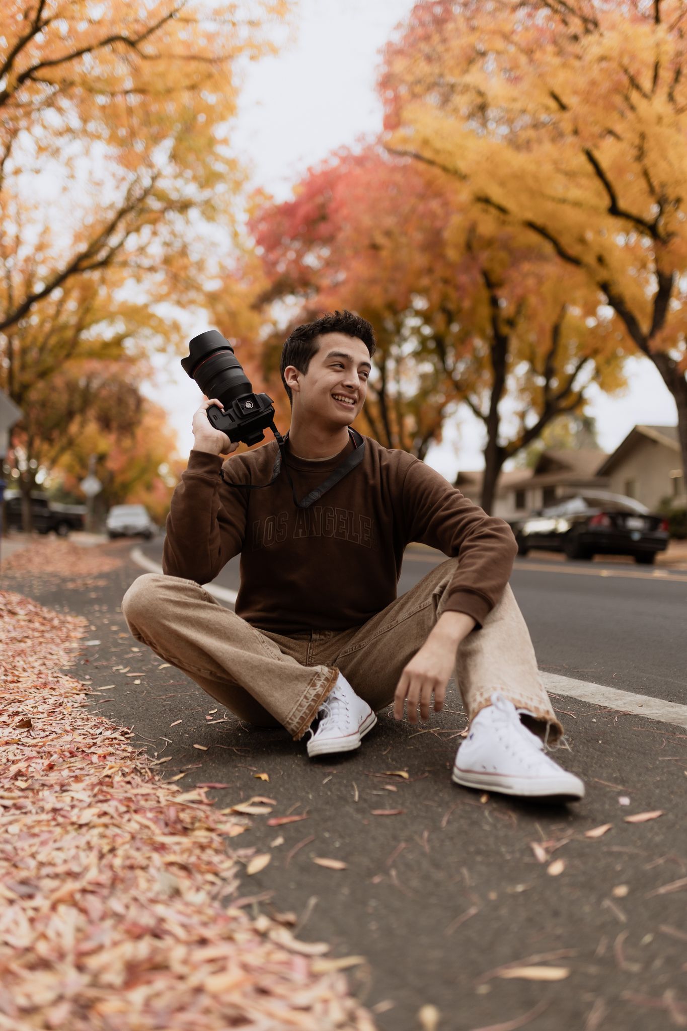 Person in brown sweater lies on pavement covered in autumn leaves while holding camera during fall photoshoot.